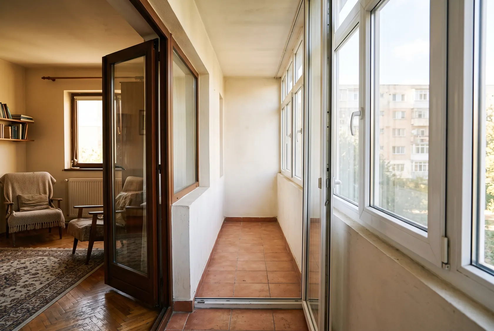 Balcon de bloc lung și îngust, închis cu tâmplărie albă, văzut din living prin ușa deschisă, cu pardoseala goală