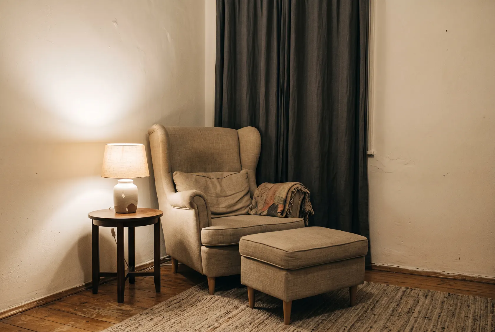 Colț pentru hrănit cu fotoliu cu spătar înalt și suport de picioare, măsuță cu lampă cu abajur textil și draperie opacă trasă