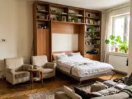 Garsonieră cu pat rabatabil vertical pe jumătate coborât, fotolii ușoare împinse lateral și dulapul care îmbracă patul până în tavan